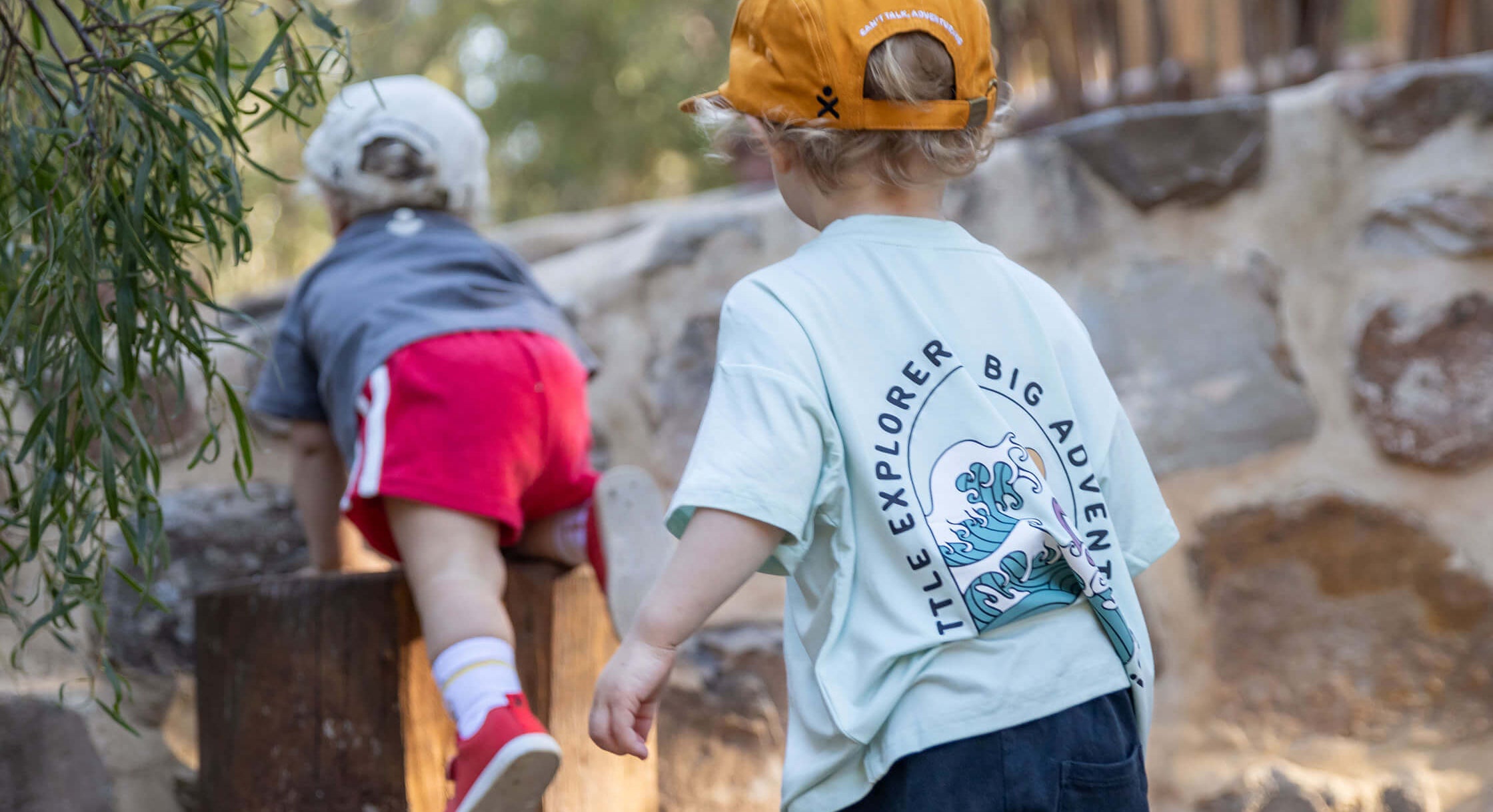 A toddler in a light blue t-shirt, navy shorts, and an orange cap stands on a wooden log at an outdoor playground, looking toward another child climbing nearby logs.