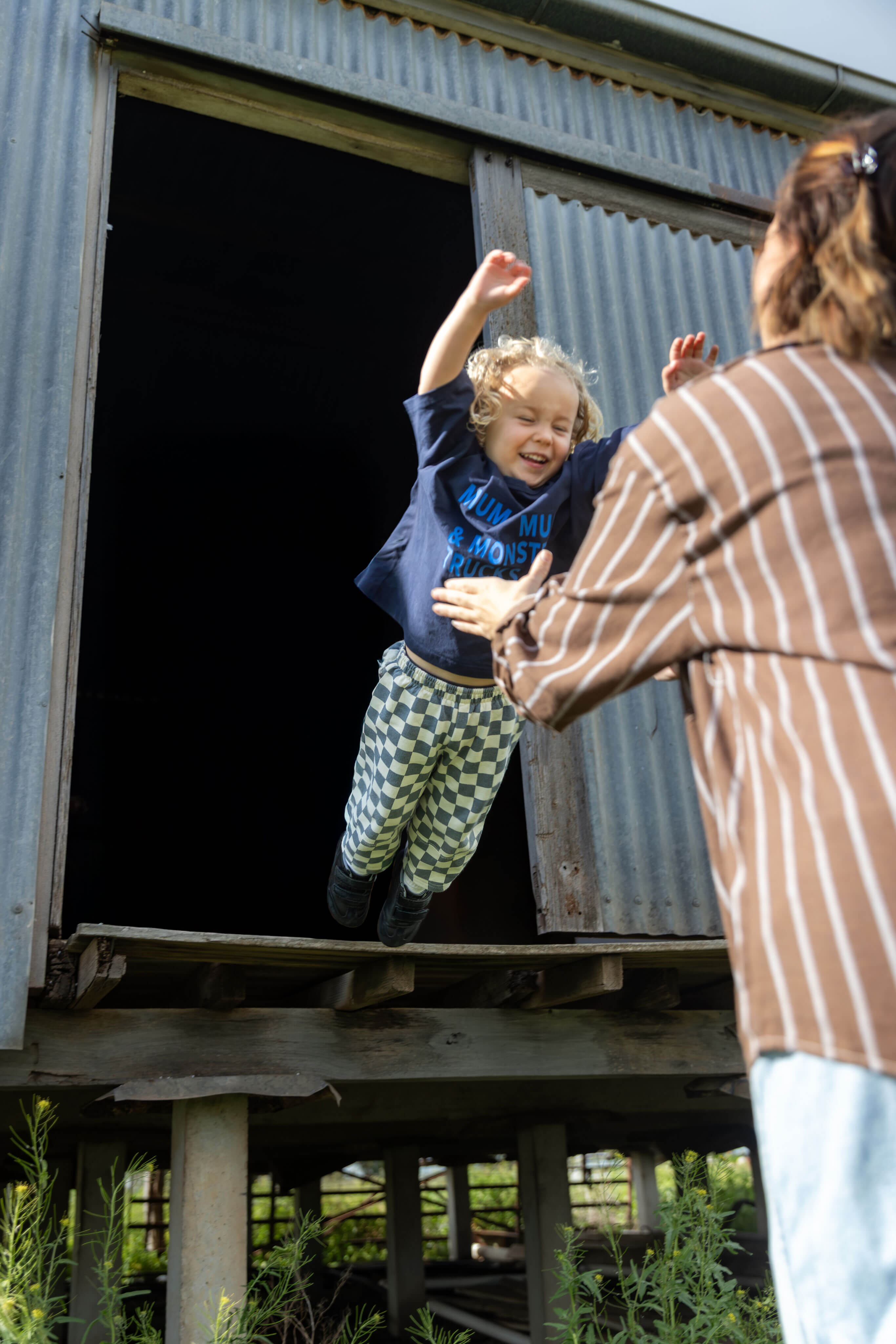 A young child wearing a navy blue t-shirt and green-and-white checkered pants jumps excitedly out of a corrugated metal elevated shed, reaching toward an adult in a brown and white striped shirt standing outside.