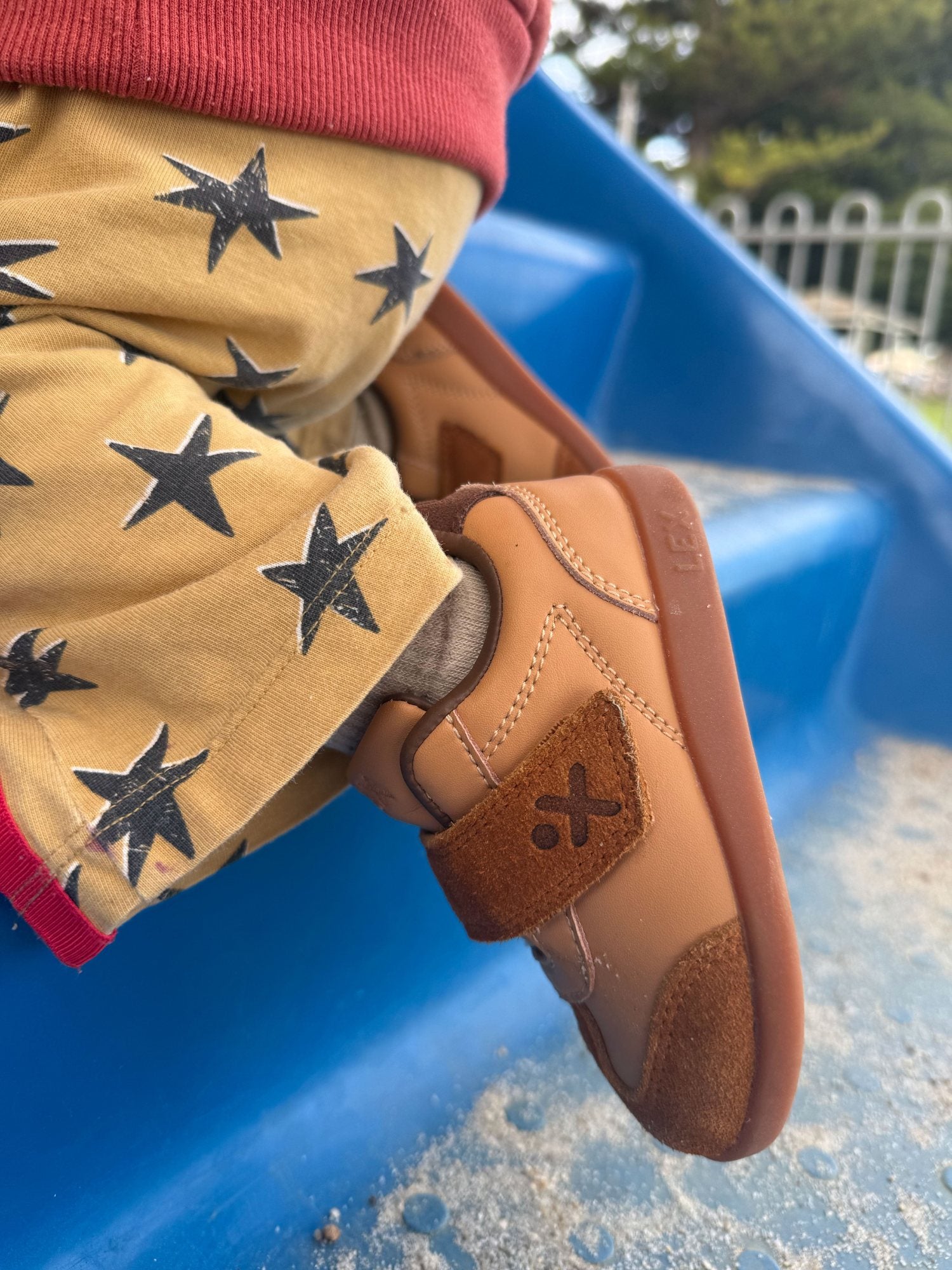 A close-up of a child's foot wearing a tan and brown sneaker with a brown strap and black star print on tan pants, sitting on a blue playground slide outdoors.
