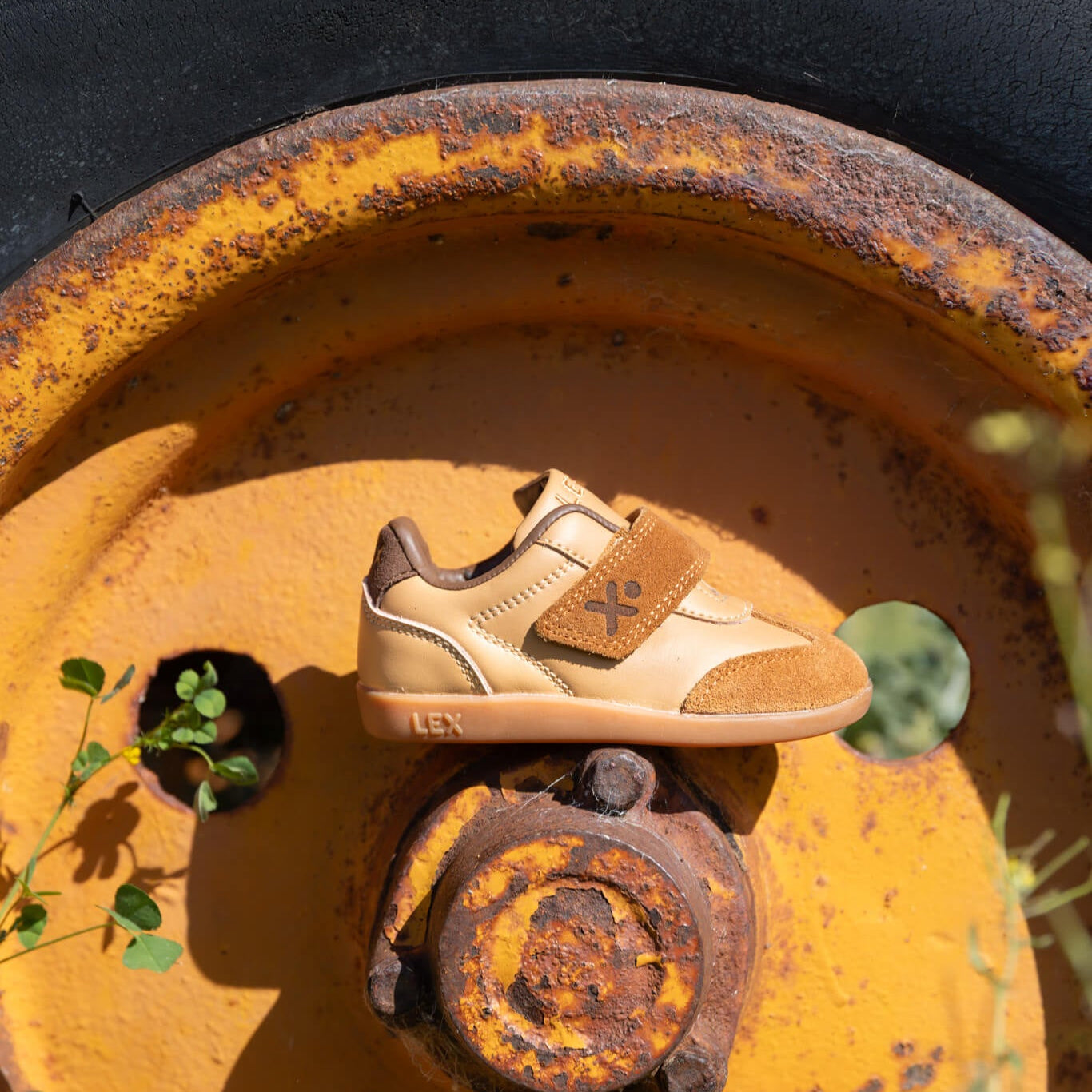 A single tan and brown low-top sneaker with a hook-and-loop strap and small logo detail, placed on a rusted yellow metal wheel hub outdoors near a tire.