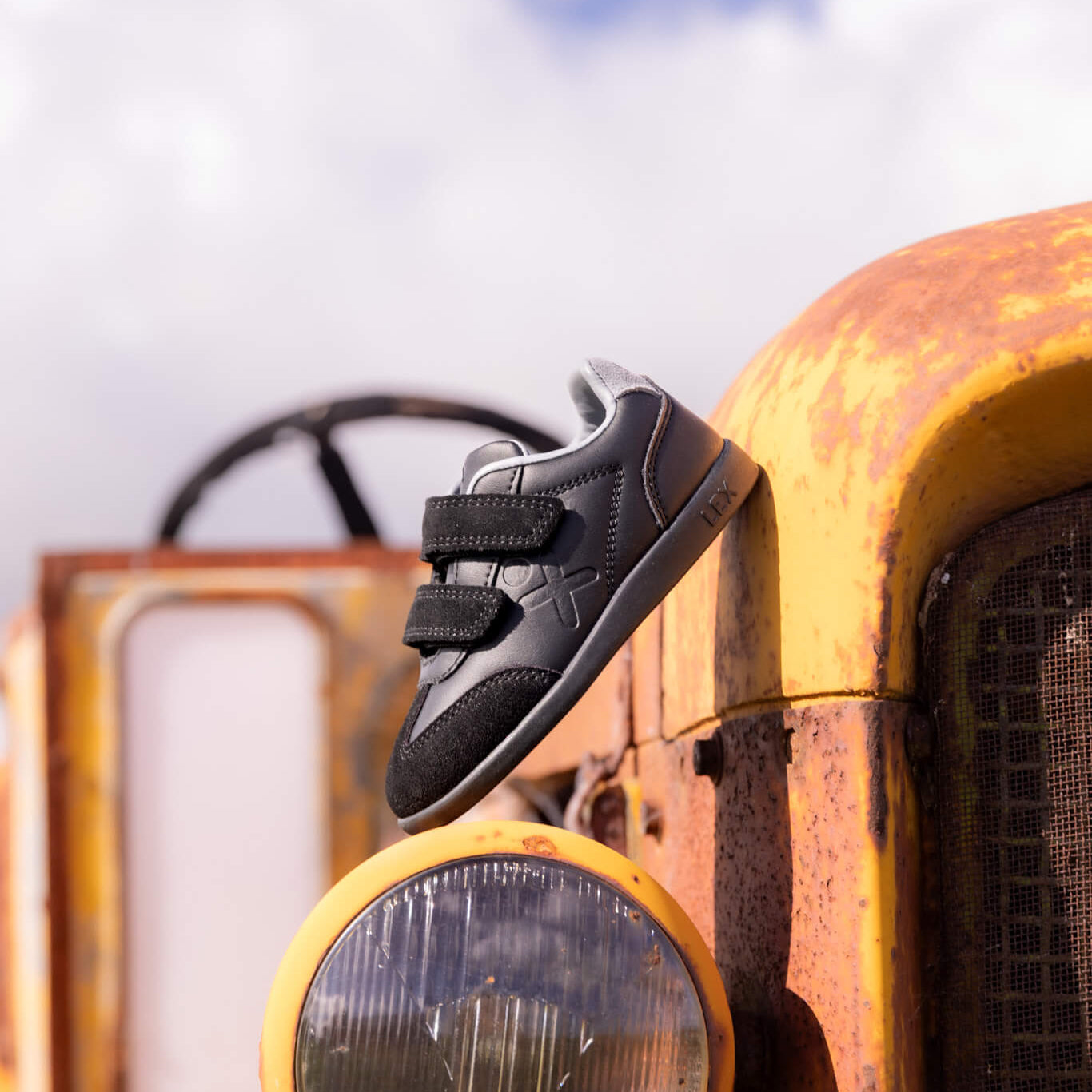 A black and gray sneaker with two hook-and-loop straps is draped over the front bumper of a rusted yellow tractor against a blue sky with clouds.