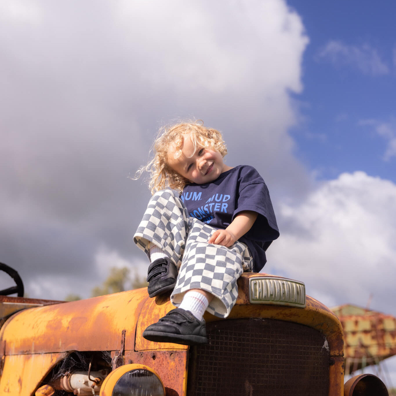 A young child with curly blonde hair sits on the rusty orange front of an old tractor outdoors under a blue sky with large white clouds. The child is wearing a navy blue t-shirt with 'RUN JUMP MONSTER' text, checkered pants, and black shoes.