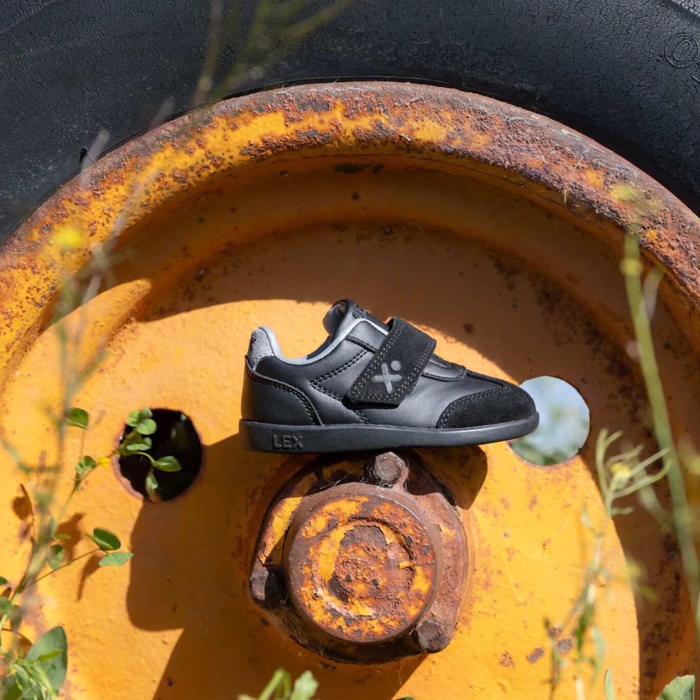 A black and gray kids' shoe with a hook-and-loop strap sits on a rusted yellow wheel outdoors, surrounded by grass and a tire.