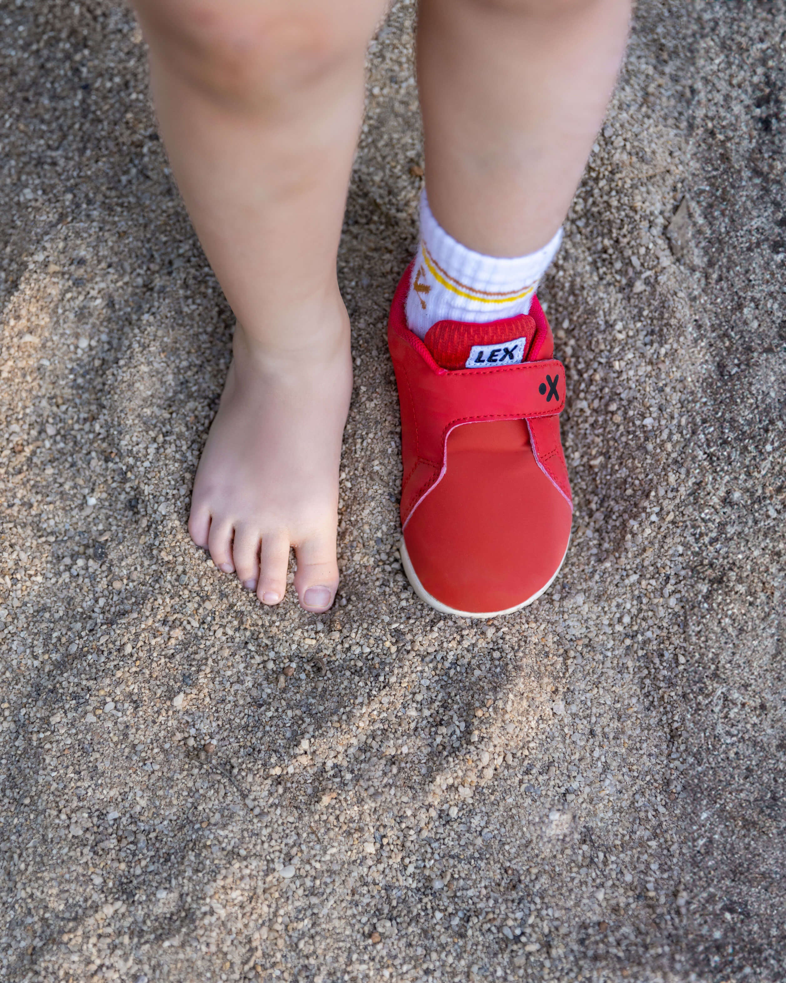 A child's bare foot and right foot wearing a red LEX slip-on shoe, standing on sandy ground.