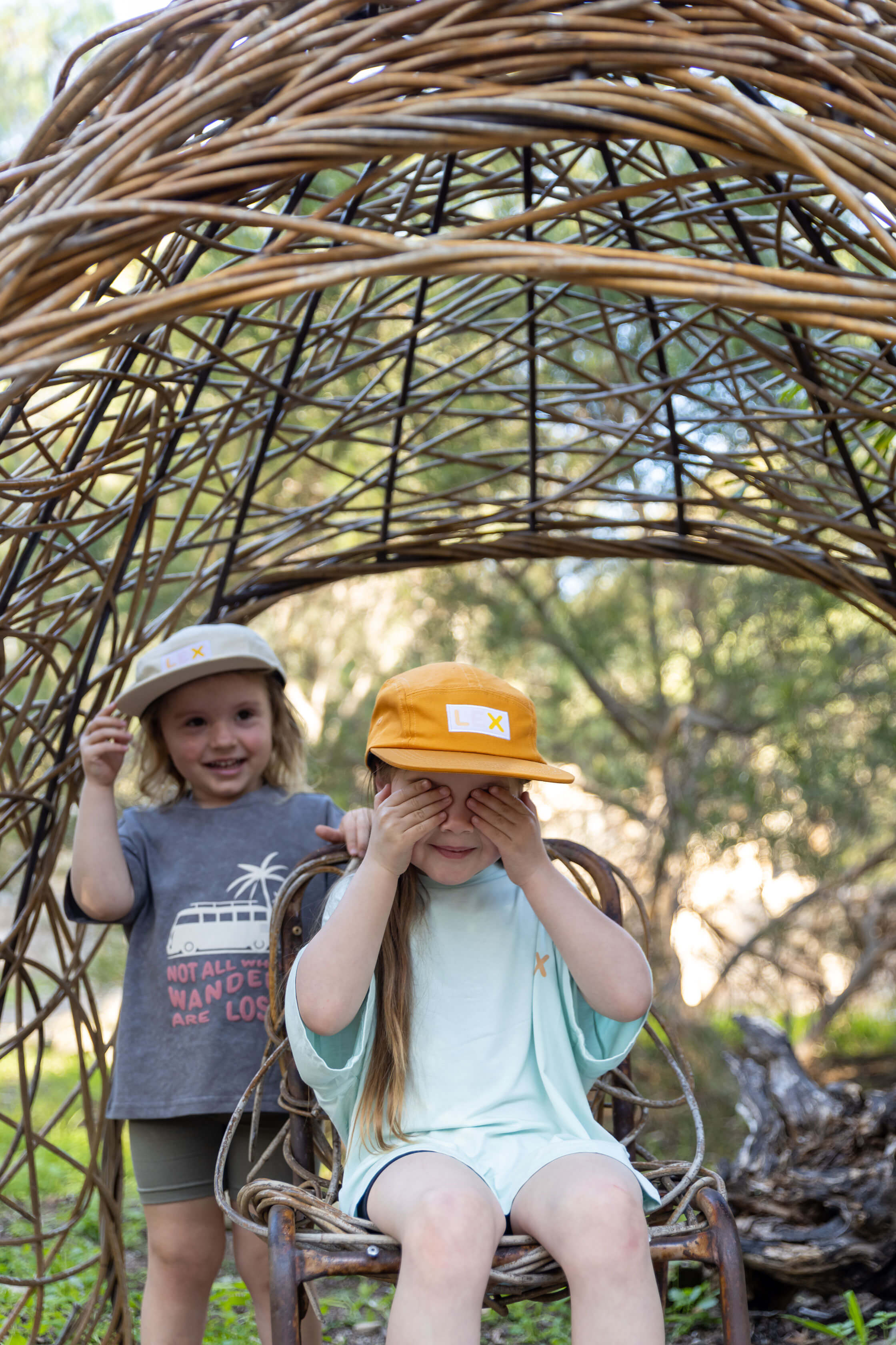 Two children outdoors under a large woven wicker arch. The child in front sits on a wicker chair wearing a yellow cap and light blue outfit, covering their eyes, while the child behind stands smiling in a gray graphic t-shirt and khaki cap.