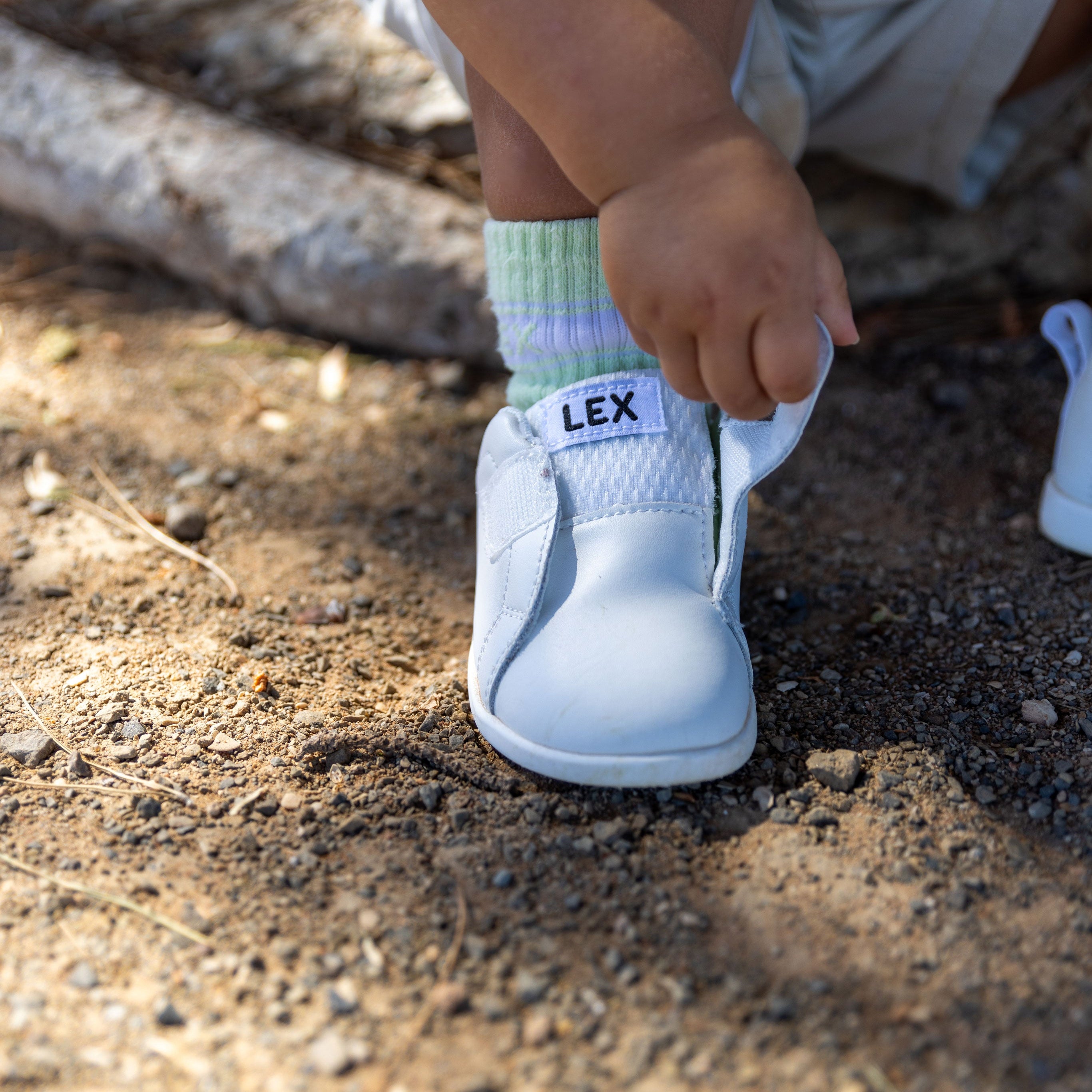 A close-up outdoor shot of a child wearing a white slip-on shoe with a green and white striped sock. The brand name 'LEX' is clearly visible on the side of the shoe.