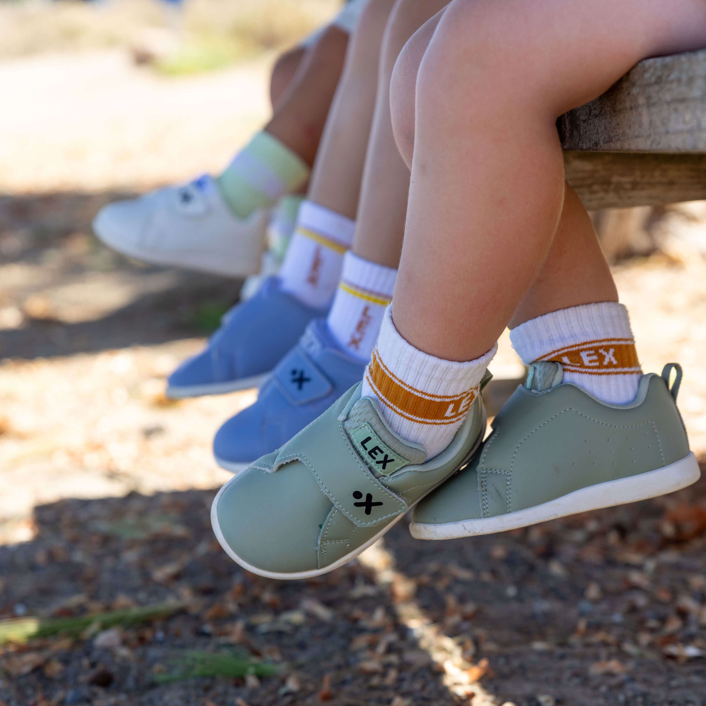 Three children's legs dangling from a bench outdoors, wearing white socks with orange bands and pastel sneakers in white, blue, and sage green. The brand name 'LEX' is visible on the socks and shoes.