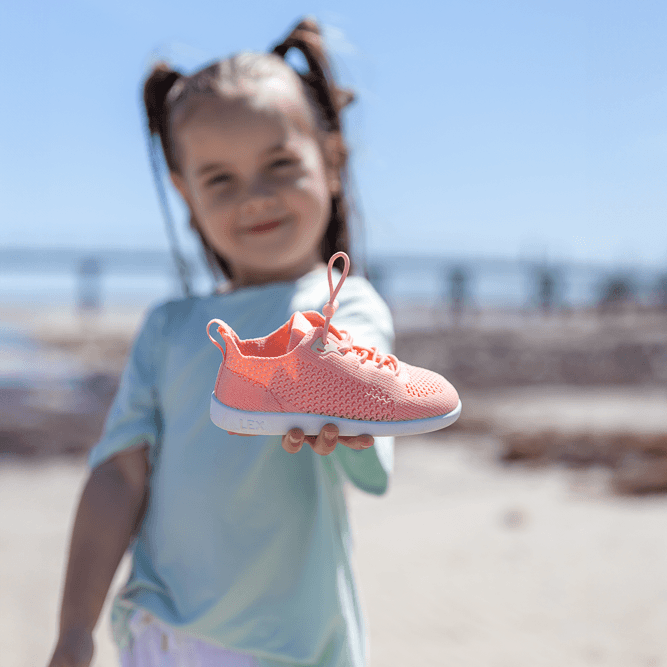 A young girl with pigtails stands on a sandy beach, holding up a coral pink athletic sneaker towards the camera. The background is a bright blue sky and the ocean, with the girl and her outfit beautifully blurred.