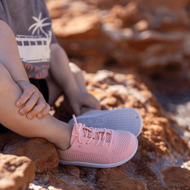 A child sitting outdoors on reddish-brown rocks wearing light pink mesh athletic sneakers with white soles.