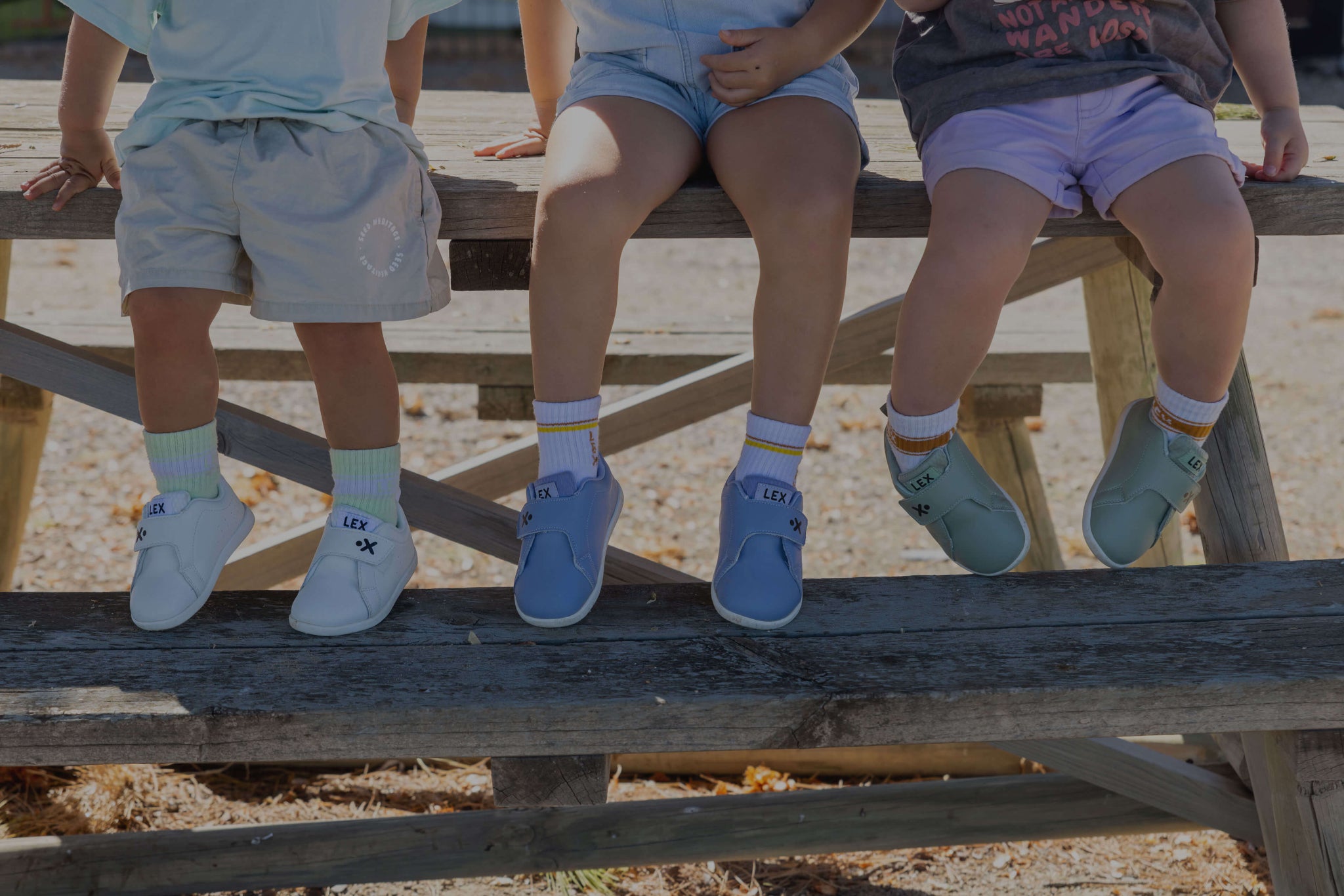 Three children sitting on a wooden picnic table outdoors, shown from the waist down wearing light blue, mint green, and gray shoes with white socks.