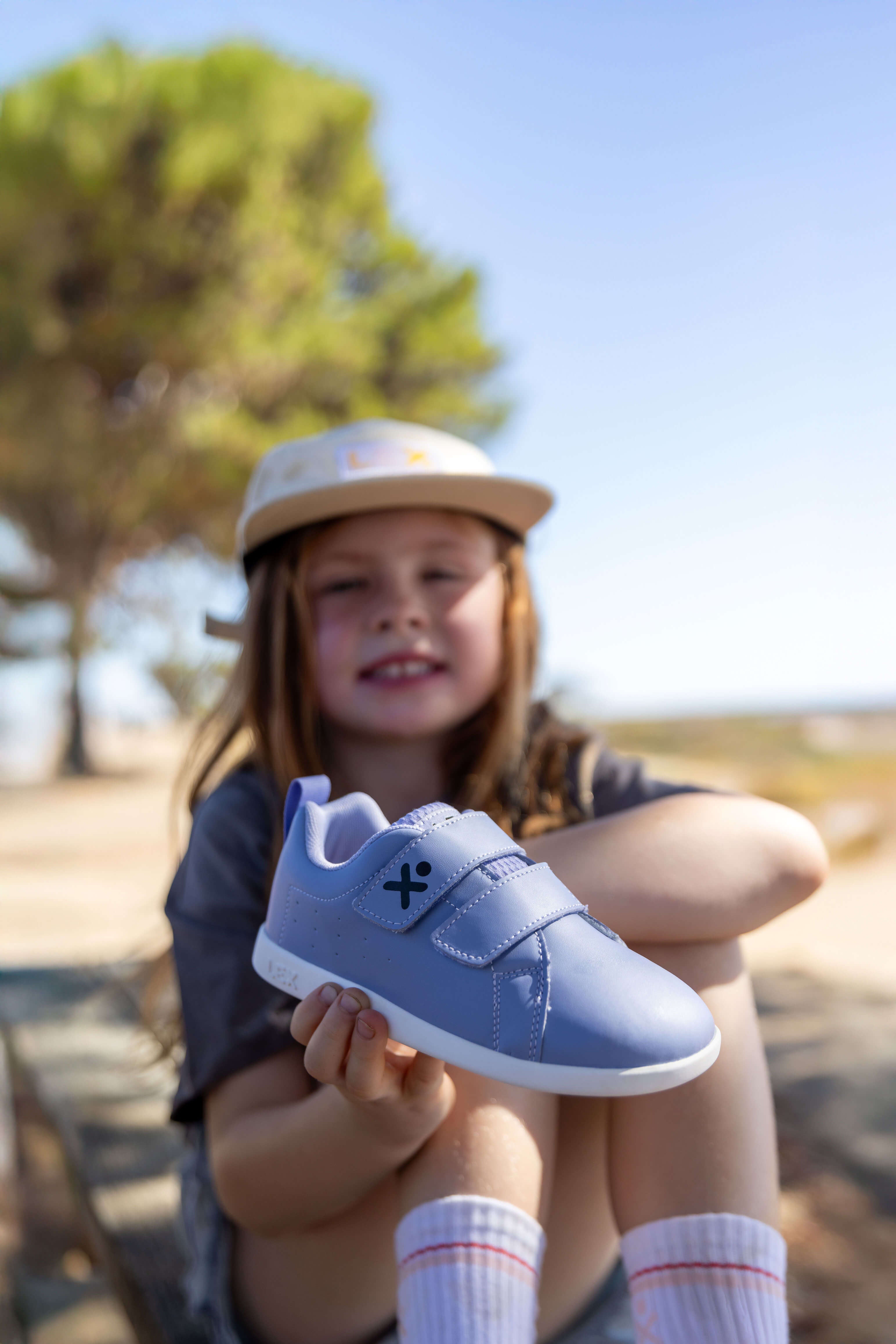 A young girl wearing a beige cap and gray shirt sits outdoors in a sunny park, holding up a light blue children's shoe with white soles. The background is a soft-focus outdoor scene with trees and blue sky.
