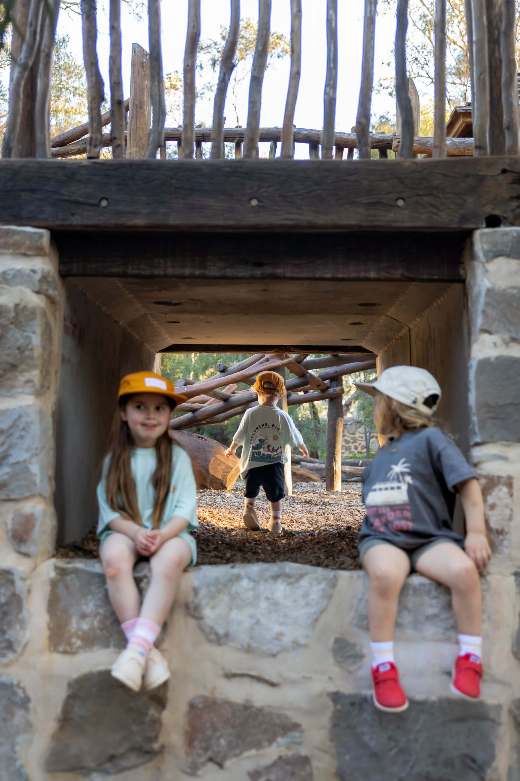 Three children playing on a rustic wooden and stone playground structure outdoors. Two children sit on the stone ledge in the foreground, and one stands in the center opening behind them.