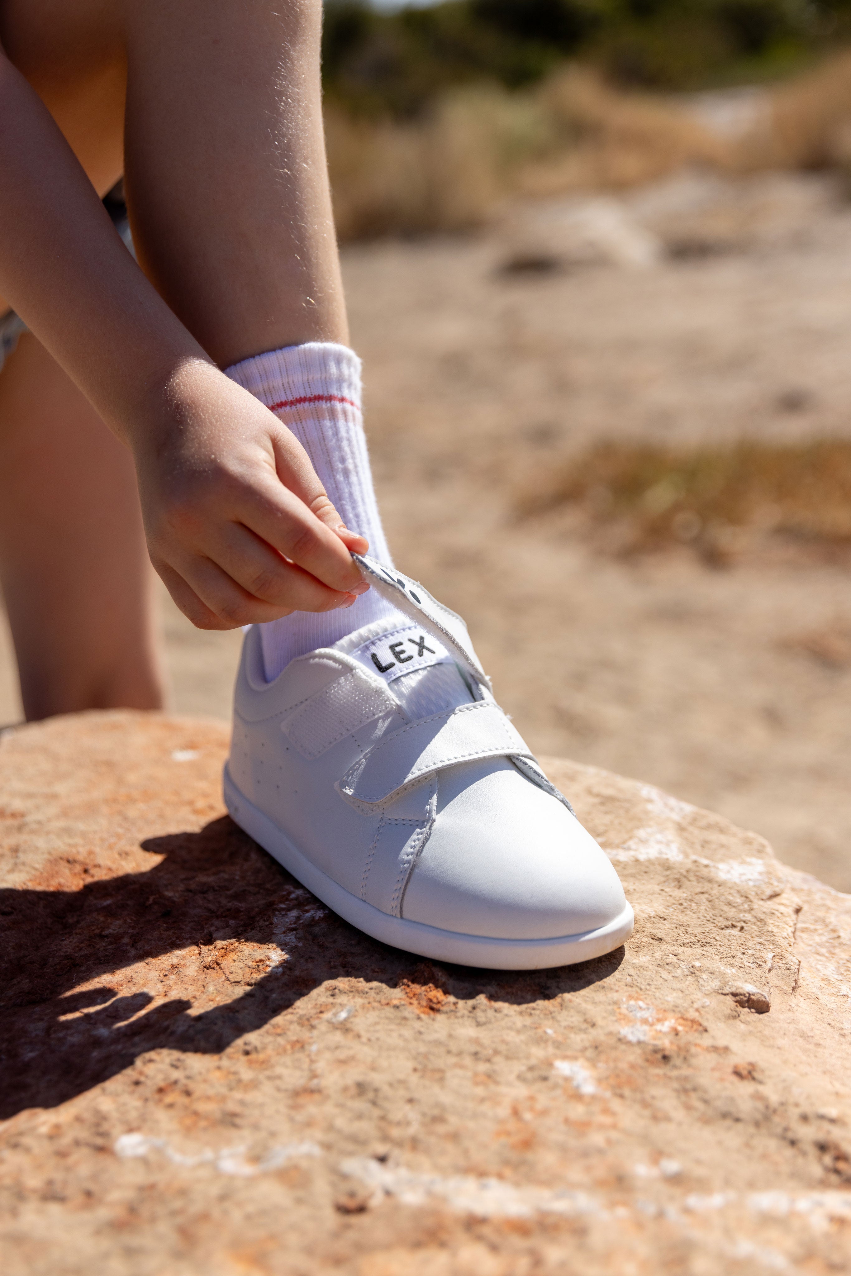 A child's white Lex sneakers with hook-and-loop straps, shown being worn outdoors on a rock with a sandy background.