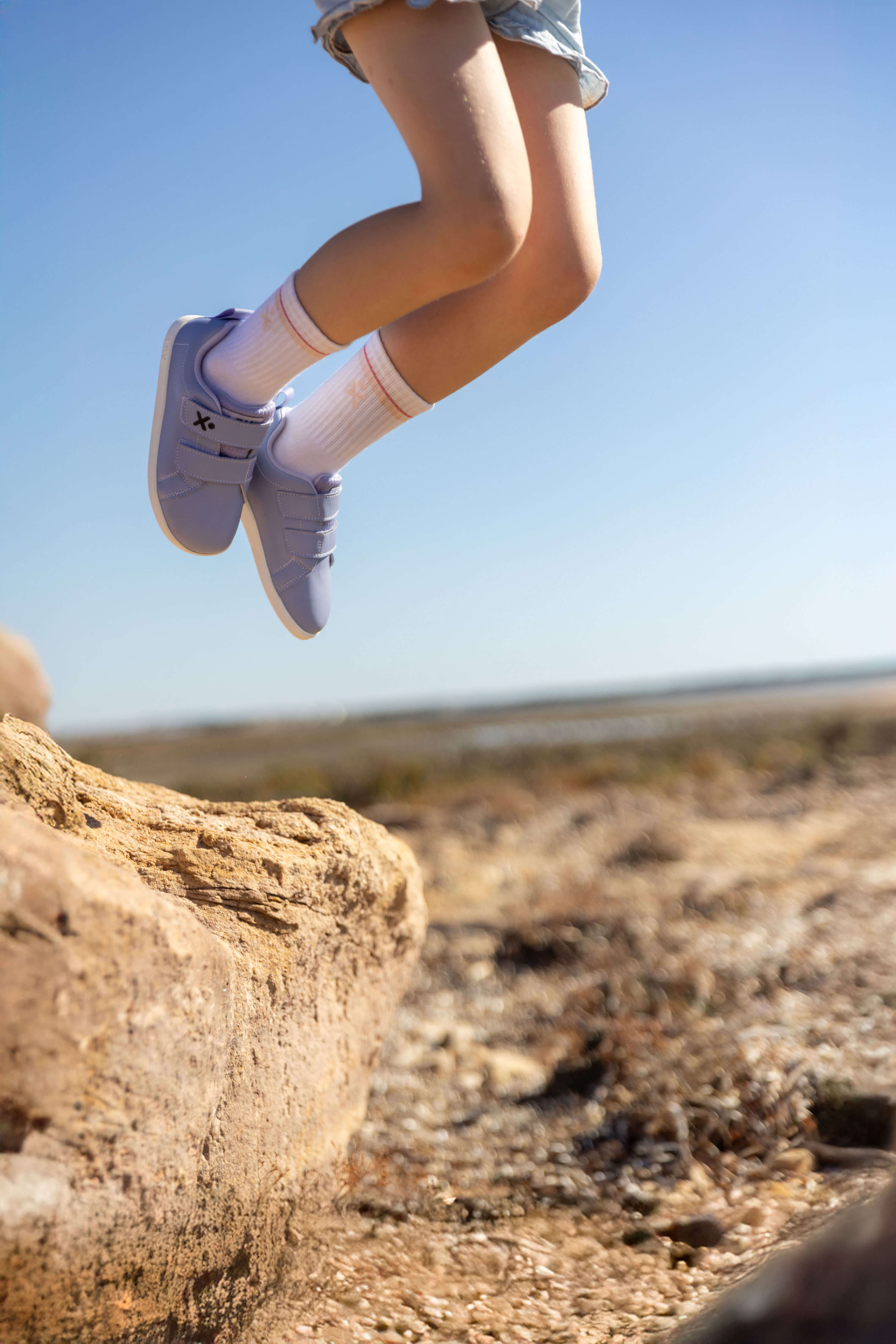 A child's legs in mid-air jumping outdoors, wearing light purple sneakers, white socks, and denim shorts against a clear blue sky and rocky desert background.