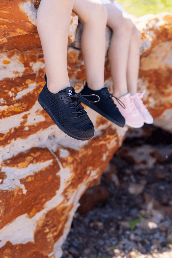 Three children's legs dangling from a rusted orange rock, wearing black and light pink sneakers against an outdoor natural background.